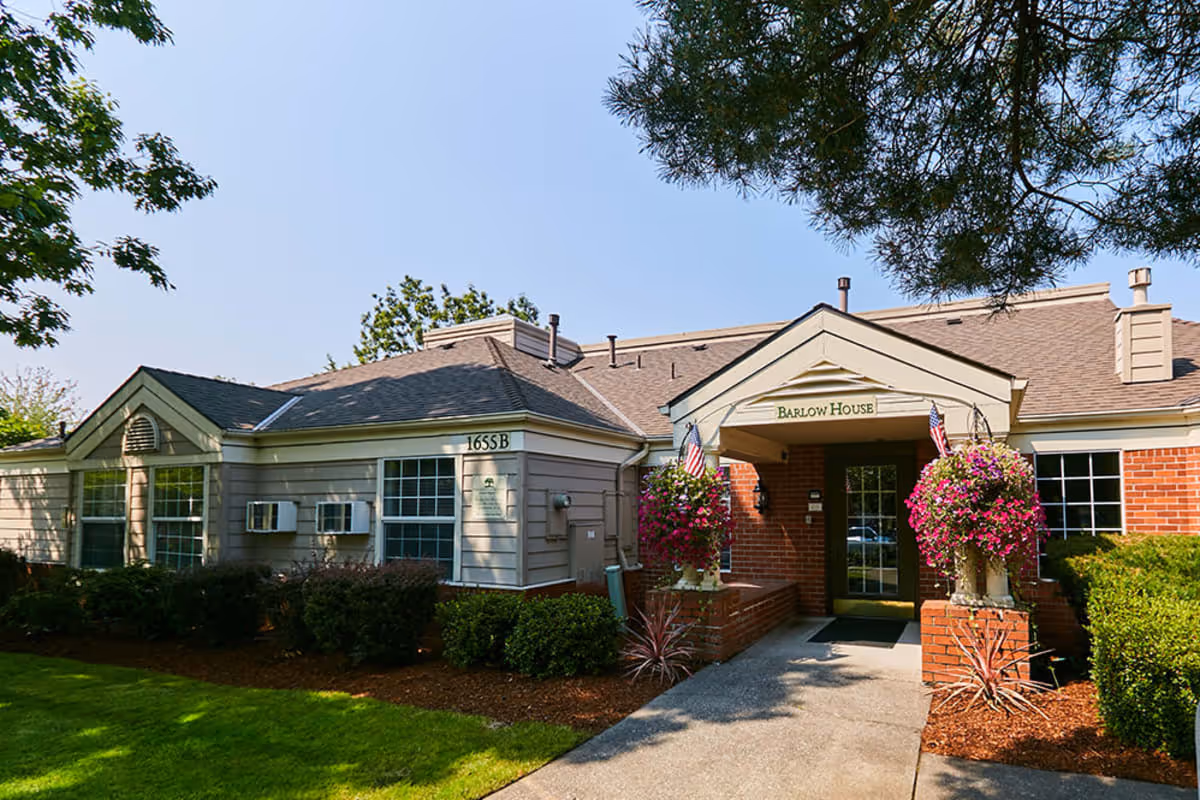 Exterior view of a single-story building named Barlow House with a brick and siding facade, surrounded by green bushes and trees, with hanging flower baskets and an American flag near the entrance.