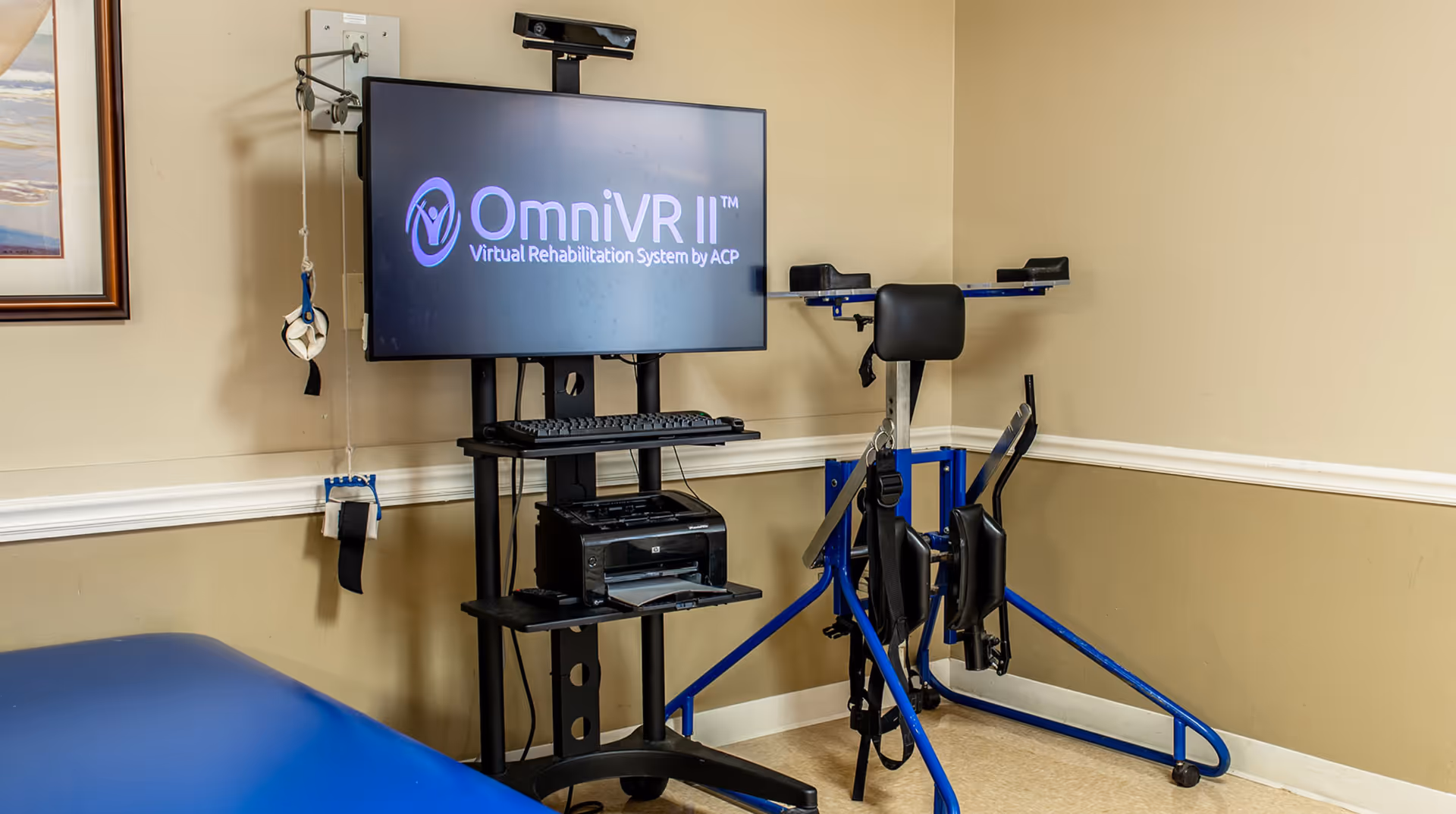 A rehabilitation room featuring a virtual rehabilitation system setup with a monitor displaying 'OmniVR II Virtual Rehabilitation System by ACP'. The setup includes a keyboard, printer, and various physical therapy equipment such as straps and supports attached to a blue metal frame. A blue therapy table is partially visible in the foreground.