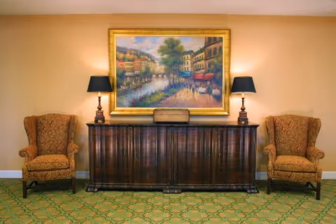 A cozy interior space featuring two patterned armchairs on either side of a dark wooden sideboard. On top of the sideboard are two matching table lamps with dark shades and a decorative box in the center. Above the sideboard hangs a large framed painting depicting a scenic riverside town with buildings, trees, and boats.
