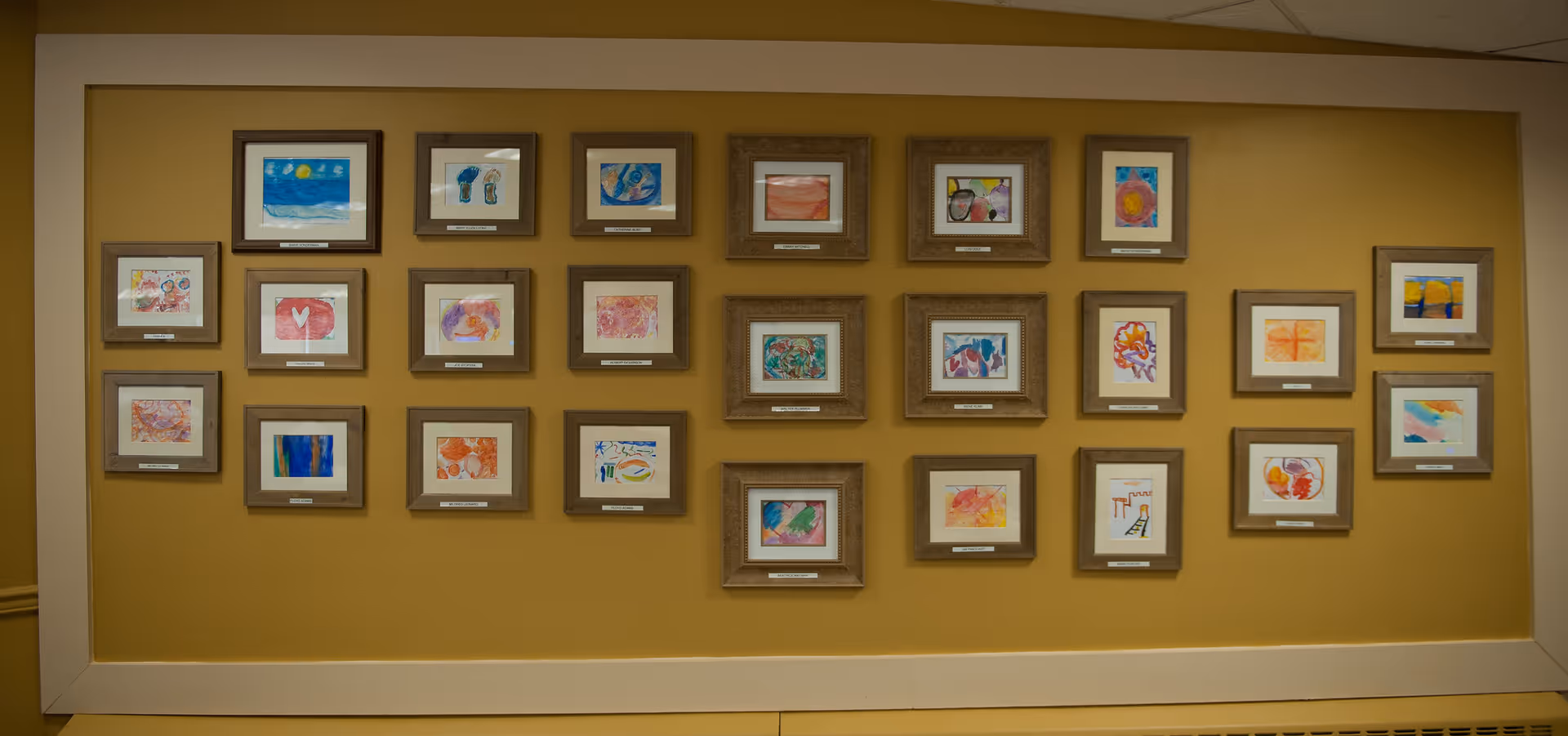 A gallery-style wall of multiple framed colorful drawings arranged in neat rows on a yellow-beige interior wall.