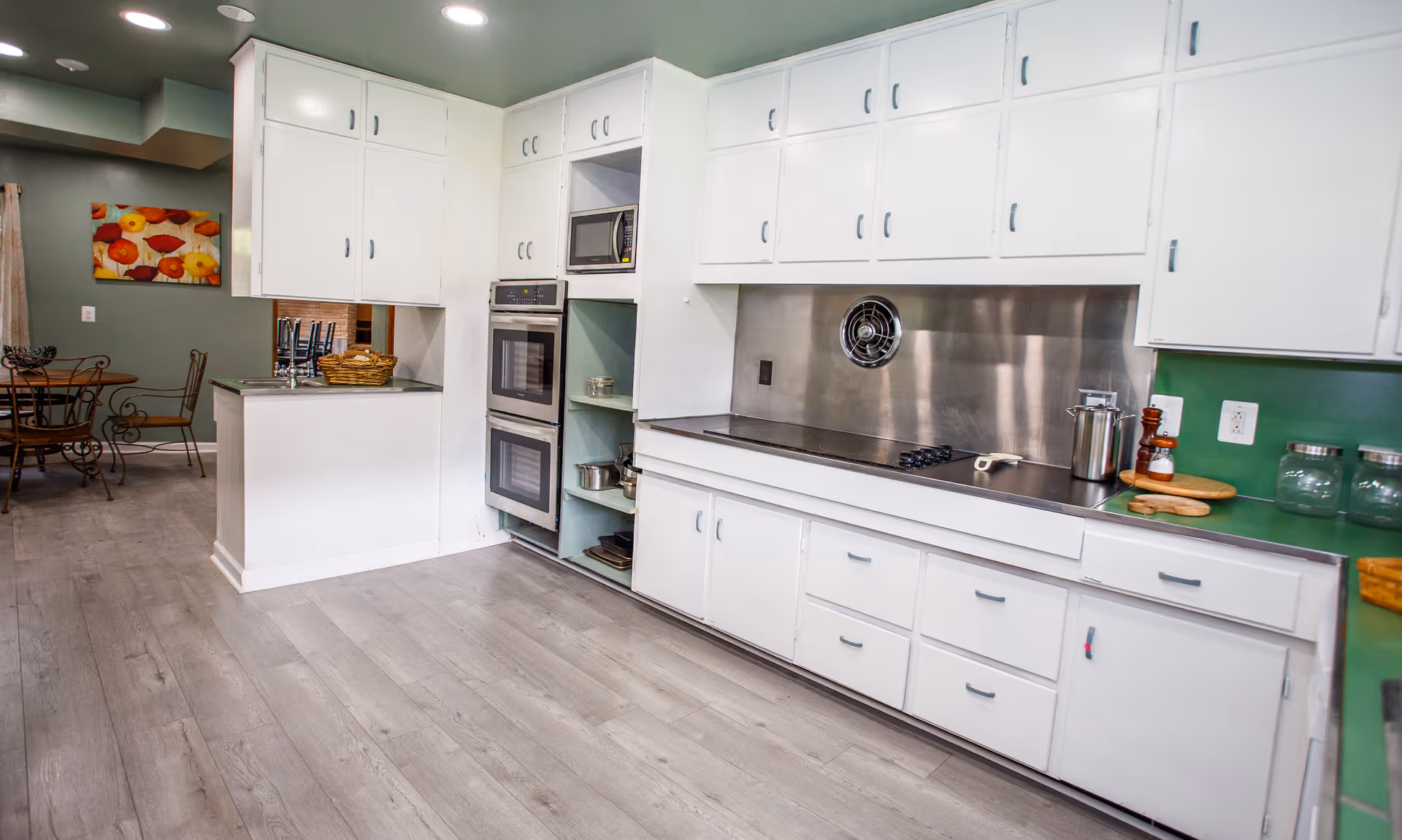Bright modern kitchen with white cabinets, stainless steel backsplash, built-in double ovens, and a small dining area visible in the background.
