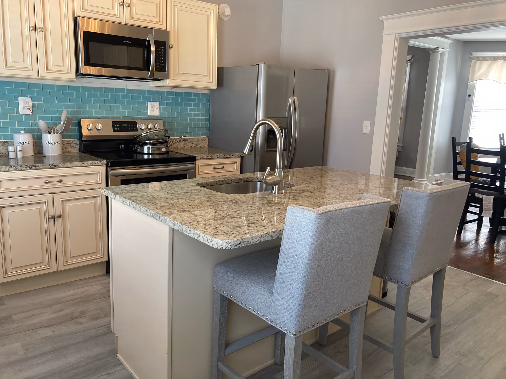 Modern kitchen with beige cabinets, stainless steel microwave, stove, and refrigerator. A kitchen island with a granite countertop and built-in sink has two gray upholstered bar stools. The backsplash is made of light blue subway tiles. In the background, a dining area with a wooden table and chairs is visible.