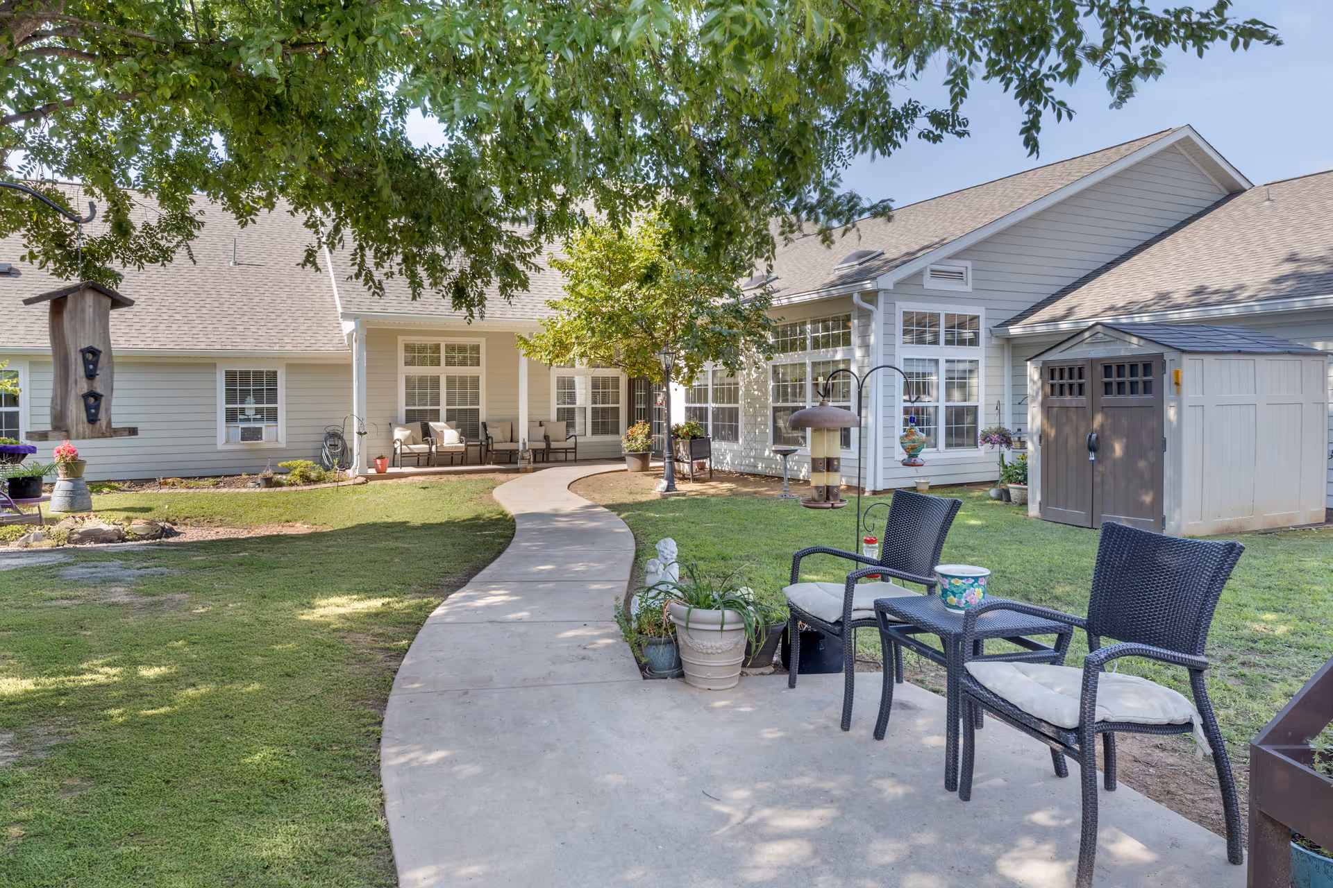 Outdoor patio area at Brookdale Norman with a curved concrete walkway leading to a building with large windows and a covered porch. There are two black wicker chairs with cushions and a small table on the right side of the walkway, potted plants, bird feeders, and a small shed. Trees provide shade over the area.
