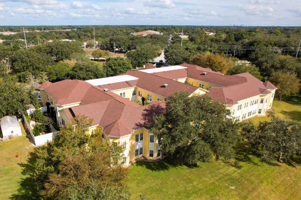 Aerial view of a large senior living facility building with yellow walls and a red roof, surrounded by green trees and grassy areas under a partly cloudy sky.