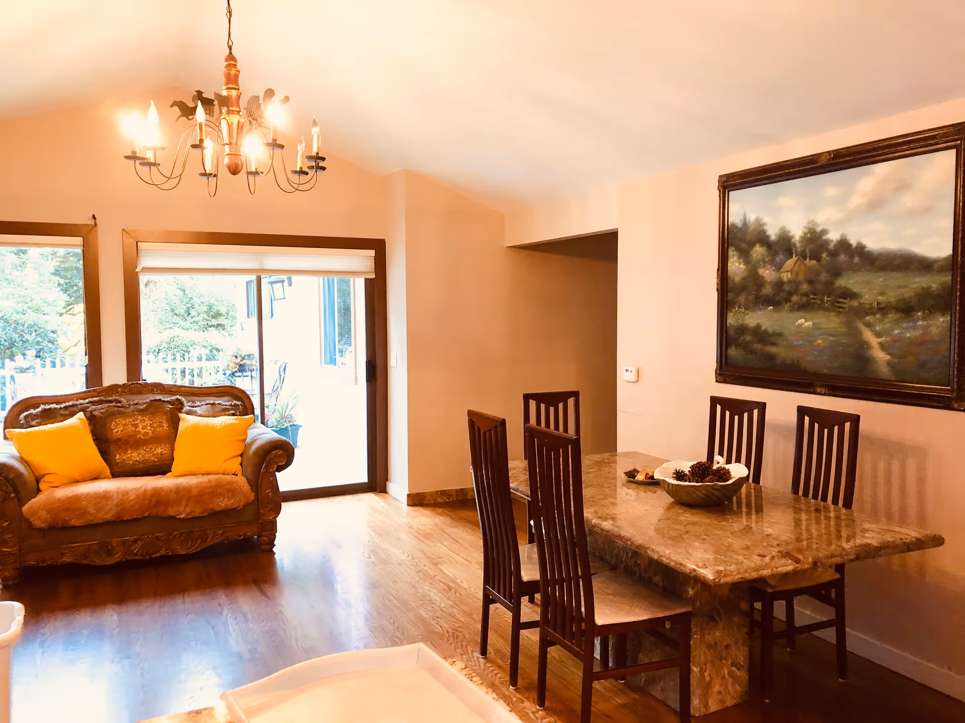 A cozy dining and sitting area with a marble dining table surrounded by six wooden chairs. A decorative bowl with pinecones is placed on the table. To the left, there is a vintage-style sofa with two yellow cushions and a brown throw. A large painting depicting a countryside scene hangs on the wall above the dining table. A chandelier with multiple lights hangs from the ceiling. Sliding glass doors with a view of greenery outside are visible behind the sofa.