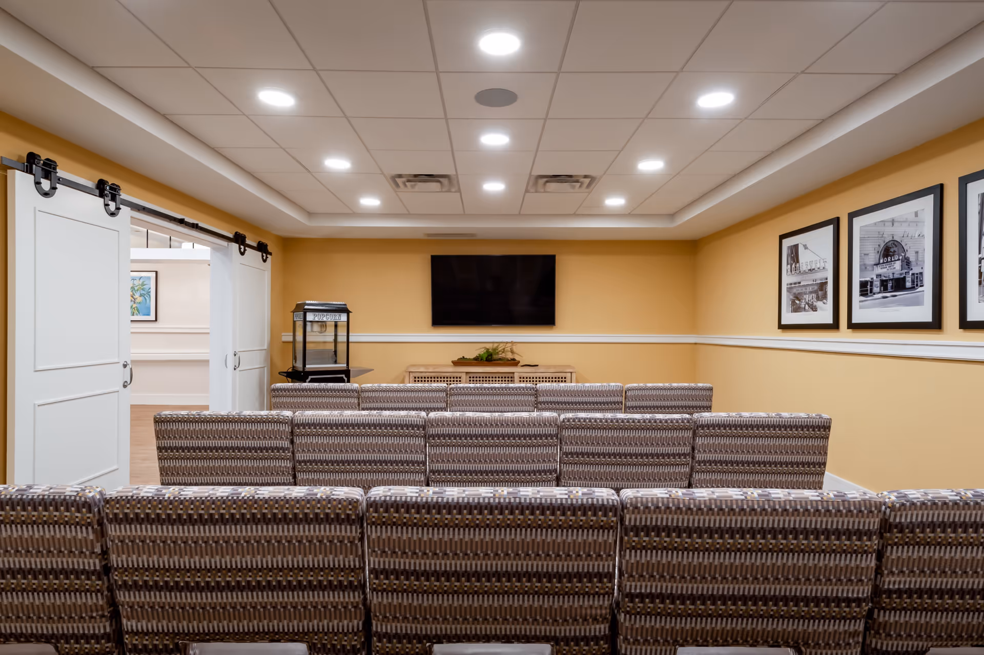 A small theater or media room with several rows of patterned chairs facing a wall-mounted flat screen TV. The walls are painted yellow with white trim, and there are three black and white framed pictures on the right wall. A popcorn machine is positioned on the left side near white sliding barn doors that lead to another room.