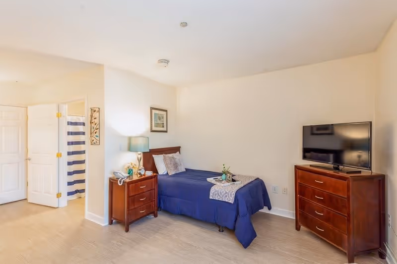 A neatly arranged bedroom in an assisted living facility featuring a single bed with blue bedding and decorative pillows. Next to the bed is a wooden nightstand with a lamp, phone, and small decor items. Across from the bed is a wooden dresser with a flat-screen TV on top. An open door reveals a bathroom with a striped shower curtain.