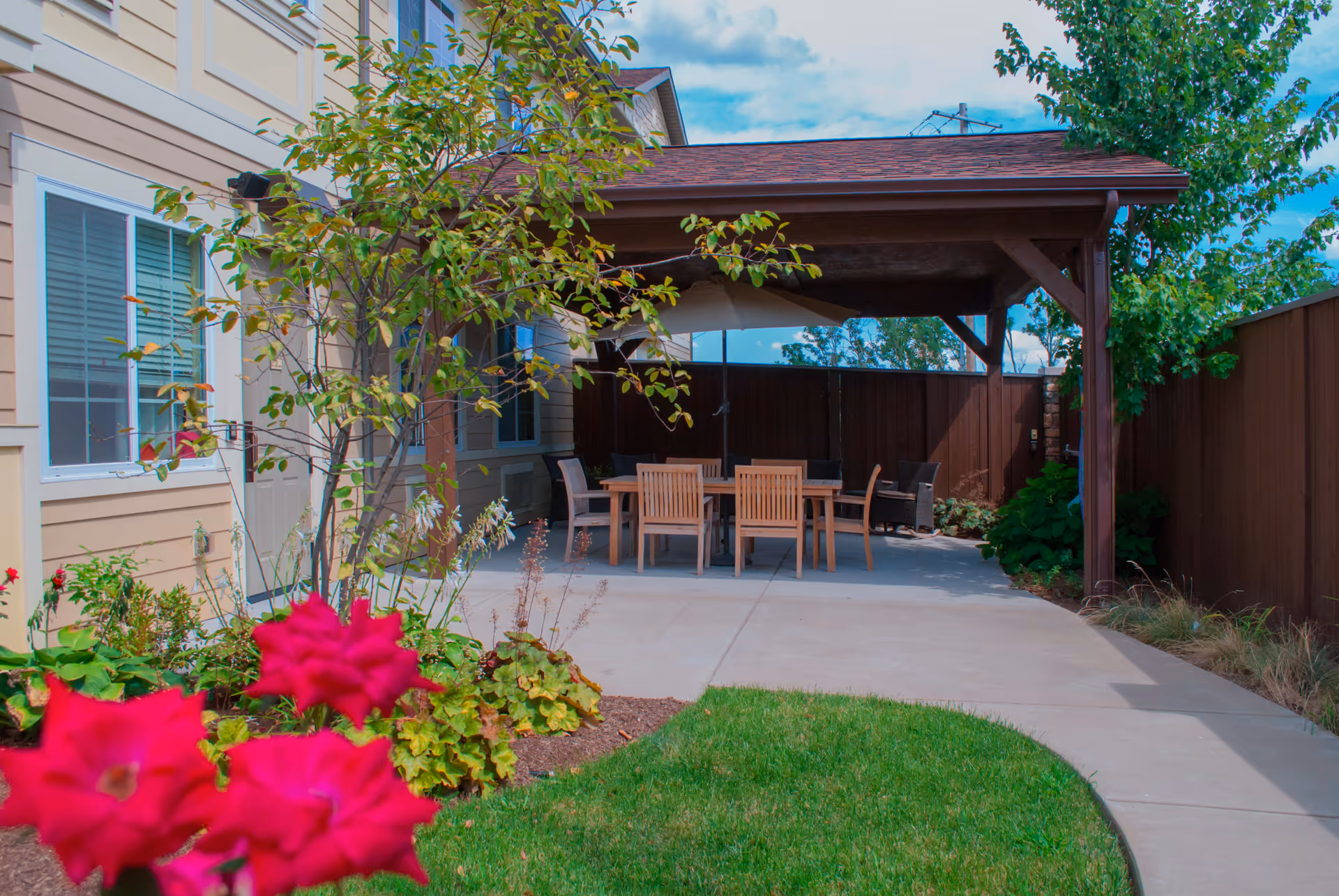 Outdoor patio area at Westview at Ellisville Assisted Living featuring a wooden pergola with a table and chairs underneath, surrounded by a garden with green plants and red flowers, adjacent to a beige building with windows.