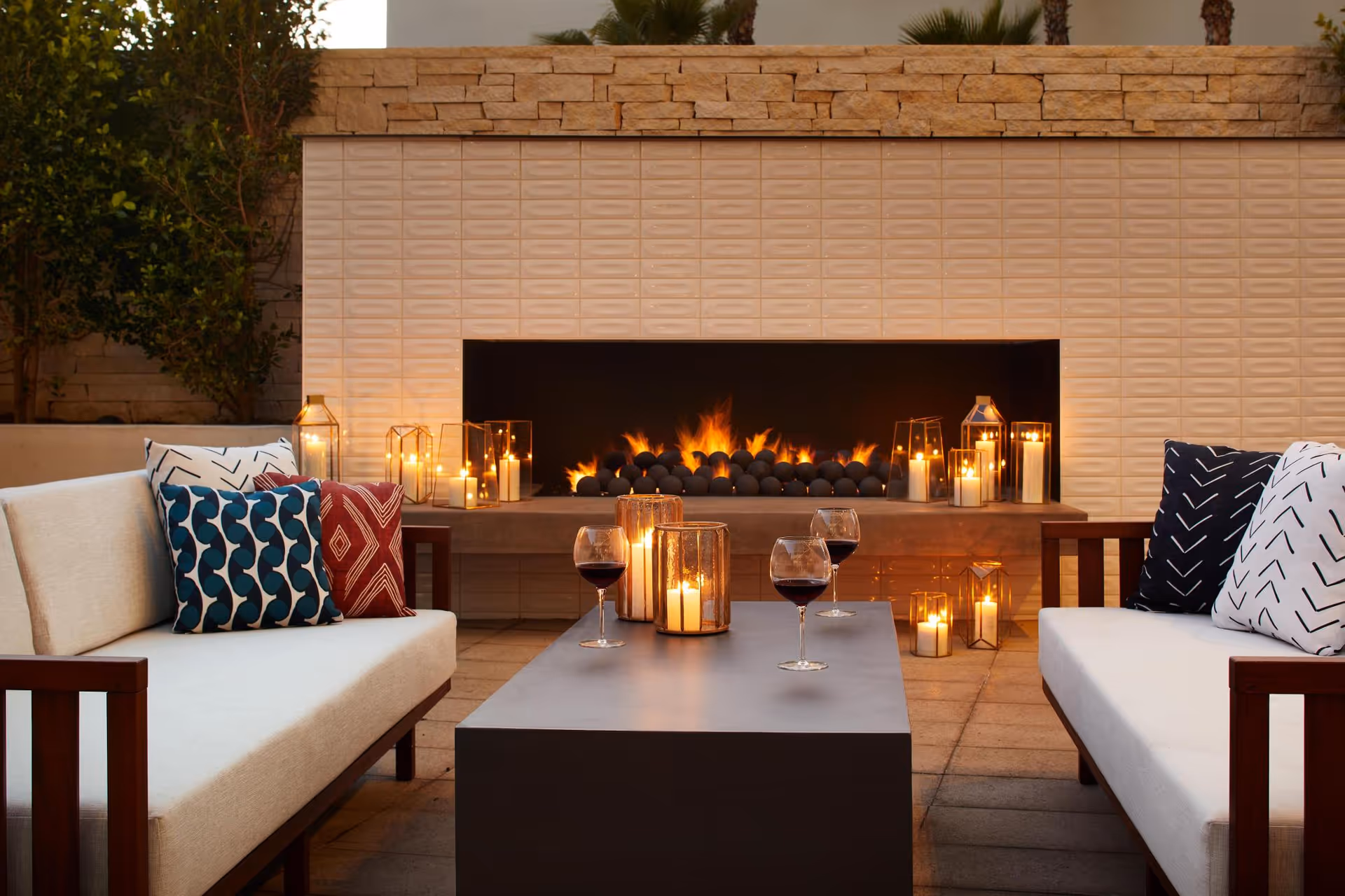Outdoor seating area with two cushioned sofas facing each other, a rectangular coffee table in between with candles and glasses of red wine, and a modern fireplace with flames and decorative lanterns in the background.