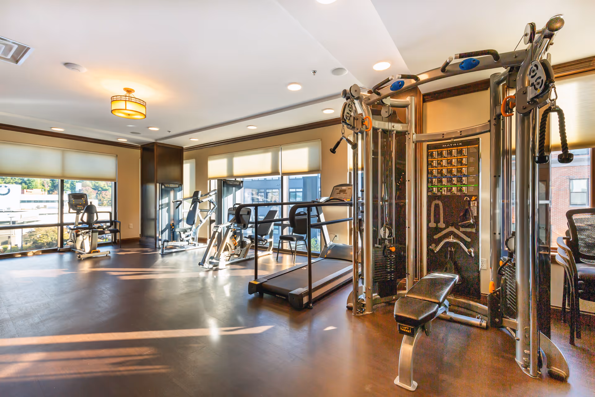 A bright fitness room with large windows allowing natural light to fill the space. The room contains various exercise equipment including a treadmill, stationary bikes, and a multi-functional weight machine. The floor is dark wood, and the walls are light-colored with dark trim. Ceiling lights provide additional illumination.