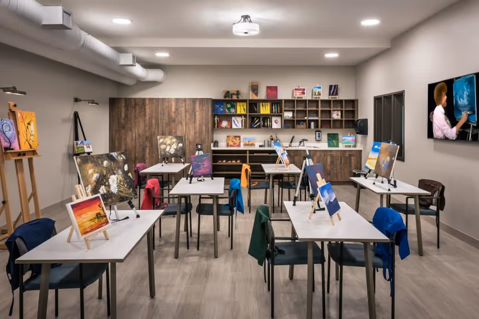 A bright art studio with several tables and chairs arranged in rows. Each table has small easels displaying colorful paintings. Jackets are draped over some chairs. The back wall features wooden cabinets and shelves filled with art supplies and more paintings. A large TV screen on the right wall shows a person painting on a canvas.