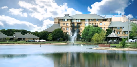 A large multi-story senior living facility building with beige exterior walls and a gray roof, situated behind a pond with a water fountain. The scene includes green trees, a gazebo near the water, and a partly cloudy blue sky.