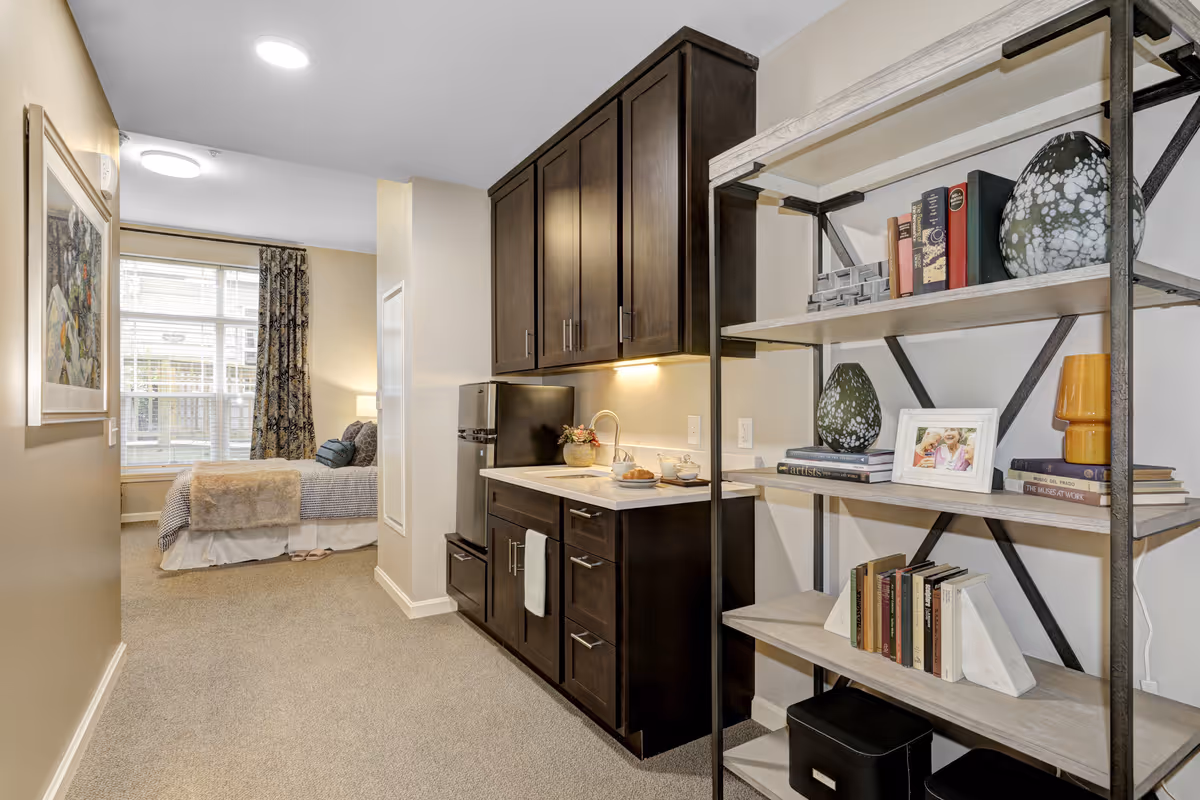Interior view of a senior living facility room showing a small kitchenette with dark wood cabinets, a mini refrigerator, and a countertop with a sink. To the right, there is a metal and wood shelving unit holding books, decorative vases, and a framed photo. In the background, a cozy bedroom area is visible with a bed, patterned curtains, and a large window letting in natural light.