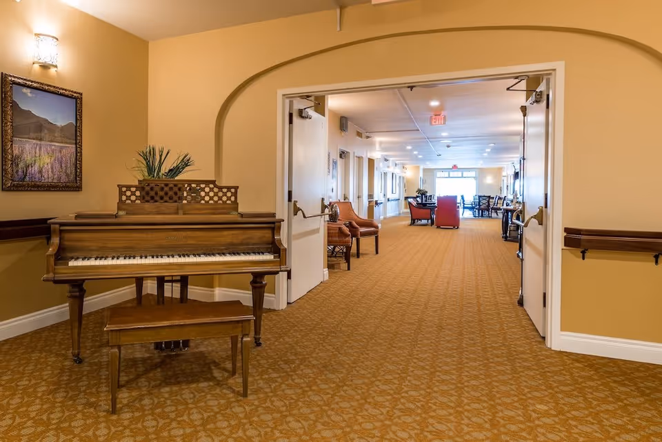 Interior view of a senior living facility hallway with a wooden piano and bench on the left side, a framed landscape picture above the piano, and an open double door leading to a well-lit common area with chairs and tables in the background.