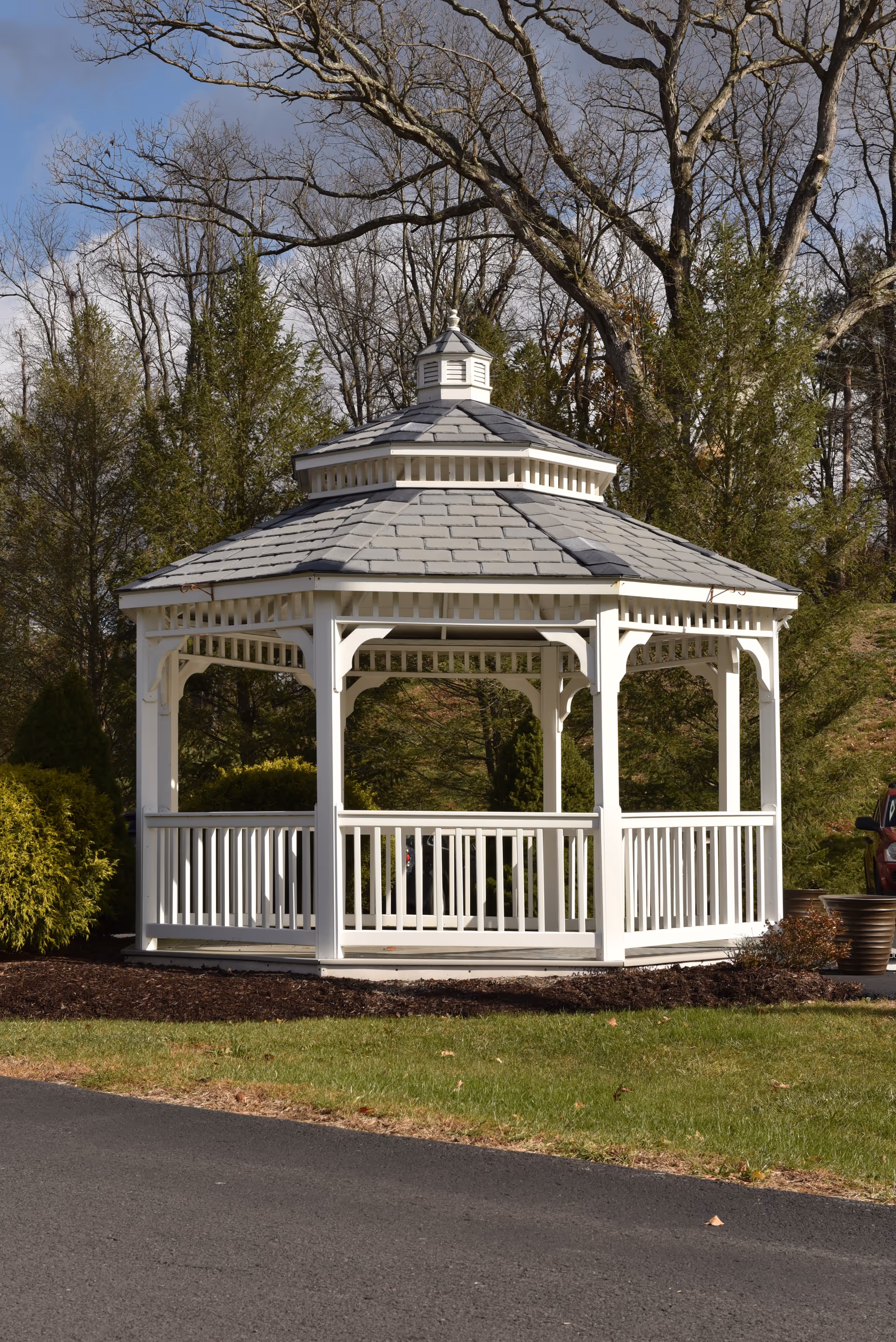 A white wooden gazebo with a gray shingled roof situated outdoors on a grassy area near a paved path, surrounded by trees and bushes under a partly cloudy sky.