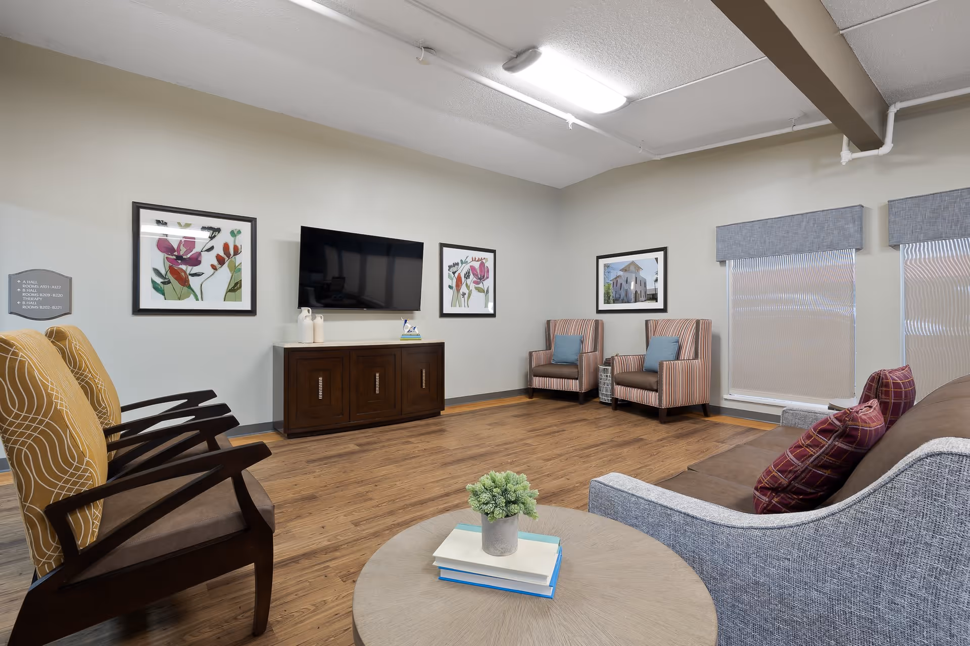 A cozy living room area in a senior living facility featuring a flat-screen TV mounted on the wall above a wooden cabinet, two striped armchairs with blue cushions, a gray sofa with red plaid pillows, and a round wooden coffee table with a small potted plant and books. The room has wooden flooring, light-colored walls with framed floral and building artwork, and two windows with blinds and valances.