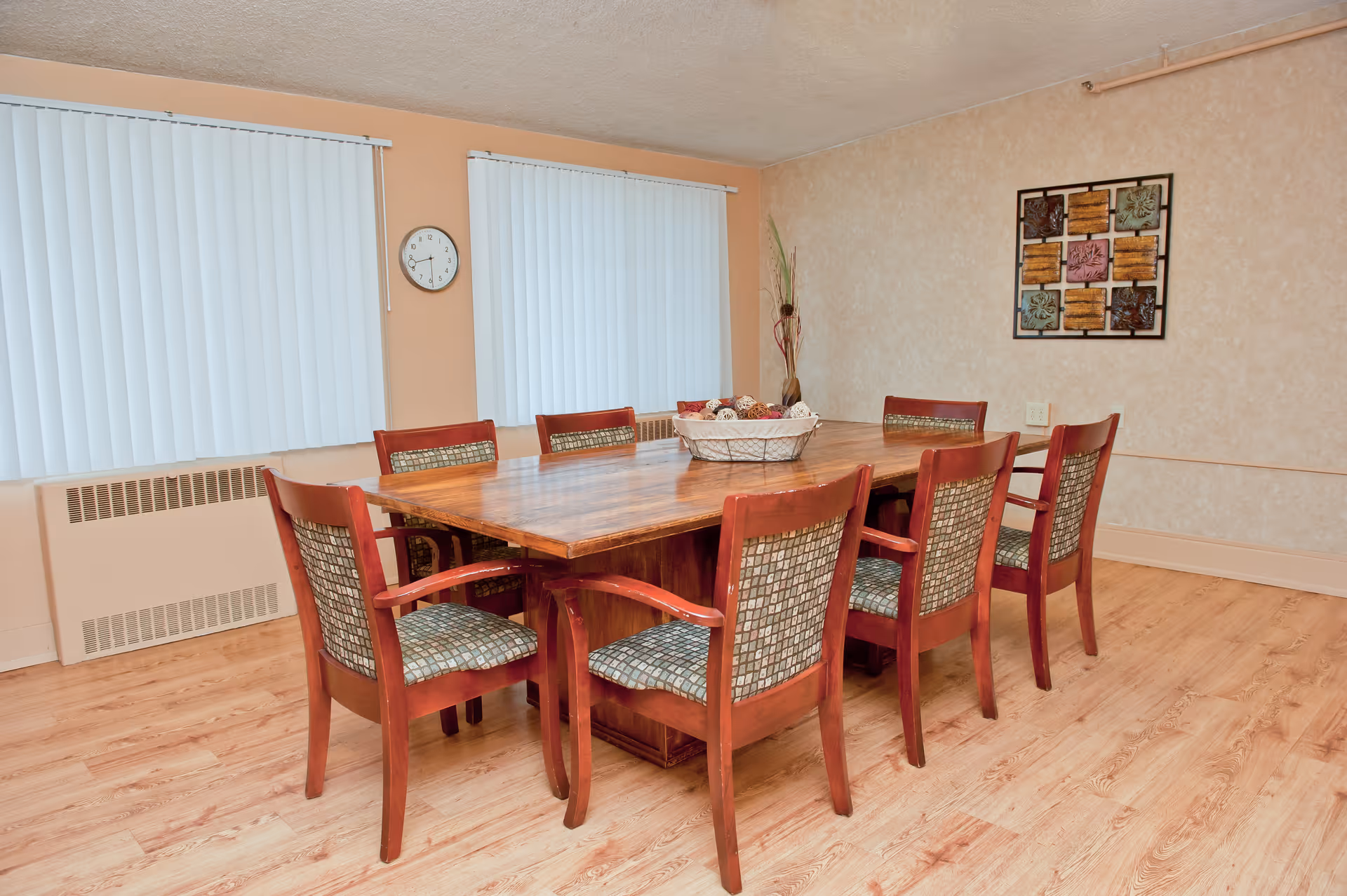 A dining room with a large wooden table surrounded by eight wooden chairs with patterned cushions. The room has light-colored walls, two windows with vertical blinds, a wall clock between the windows, a decorative wall art piece, and a basket centerpiece on the table. The floor is light wood.