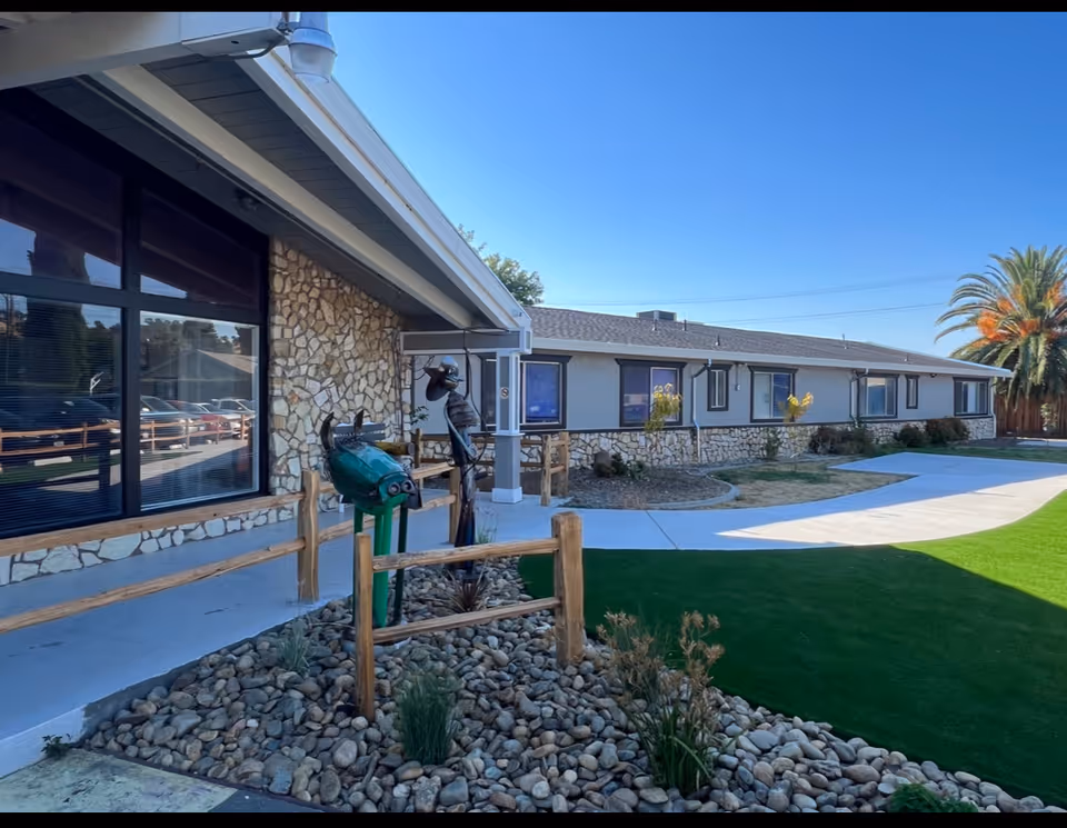 Exterior front of a single-story building with stone veneer, large windows, a paved walkway, decorative rock landscaping and artificial turf under a clear blue sky.