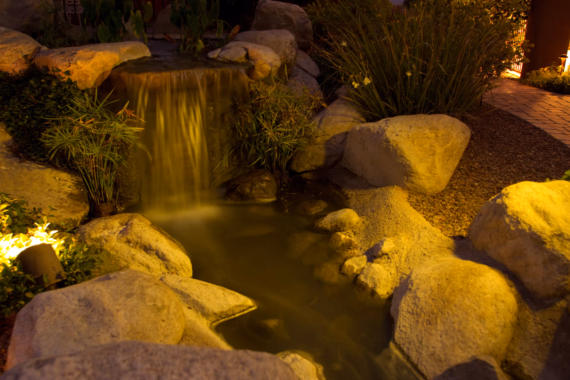 A small, illuminated outdoor waterfall feature surrounded by large rocks and plants at night, with a paved walkway visible on the right side.