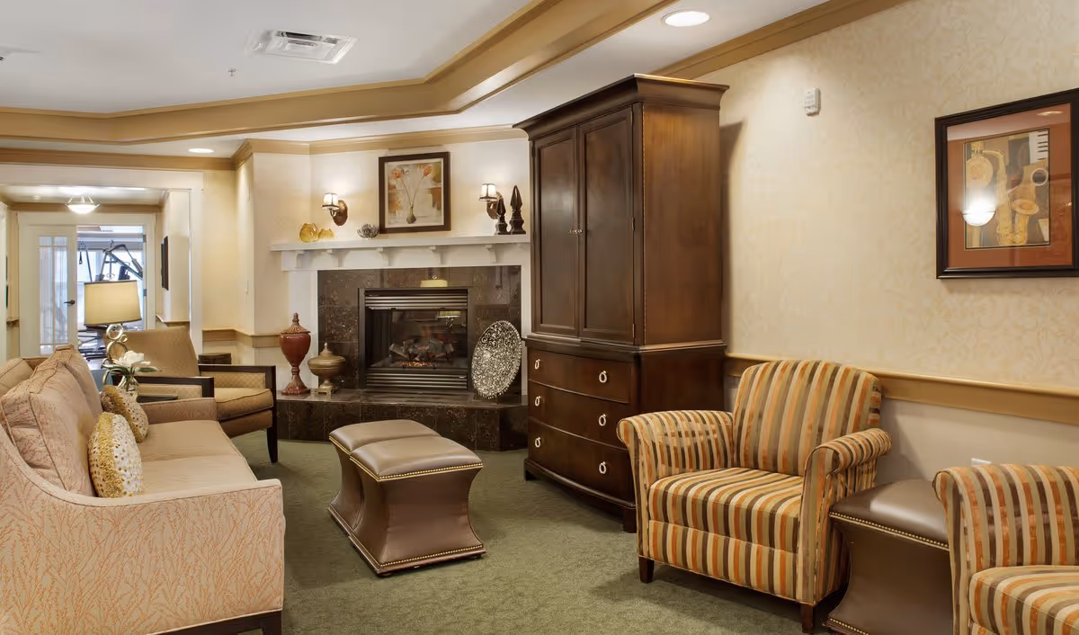 A cozy living room with a beige sofa, two striped armchairs, a dark wooden cabinet, and a fireplace with decorative items on the mantel. The room has soft lighting, framed artwork on the walls, and a green carpeted floor.