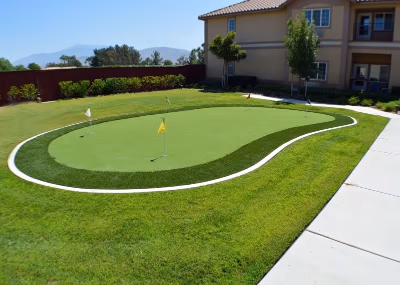 Outdoor putting green with several small flags on a well-maintained lawn next to a two-story building under a clear sky.