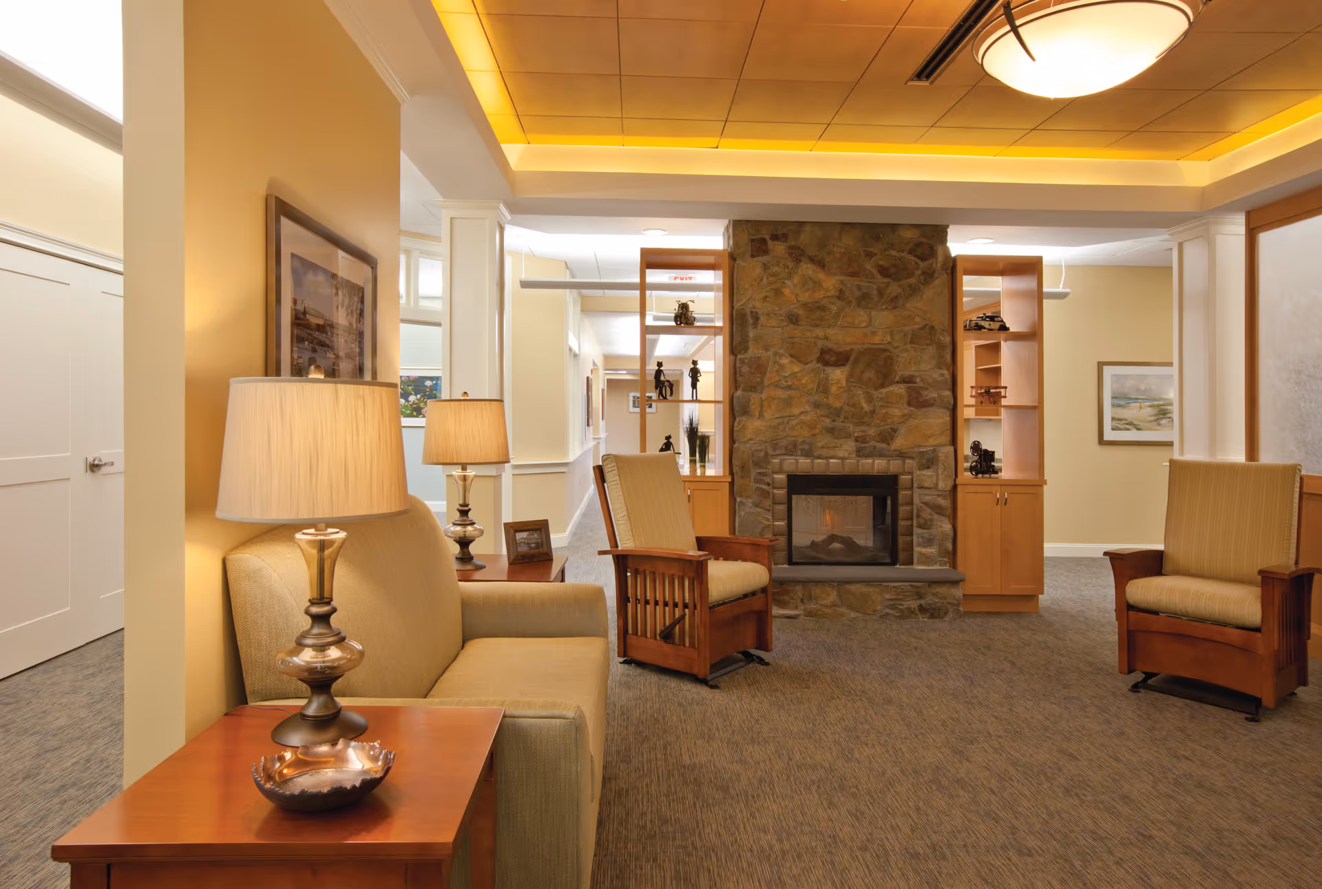Comfortable common room with upholstered chairs and a sofa arranged around a stone fireplace under warm ambient lighting.