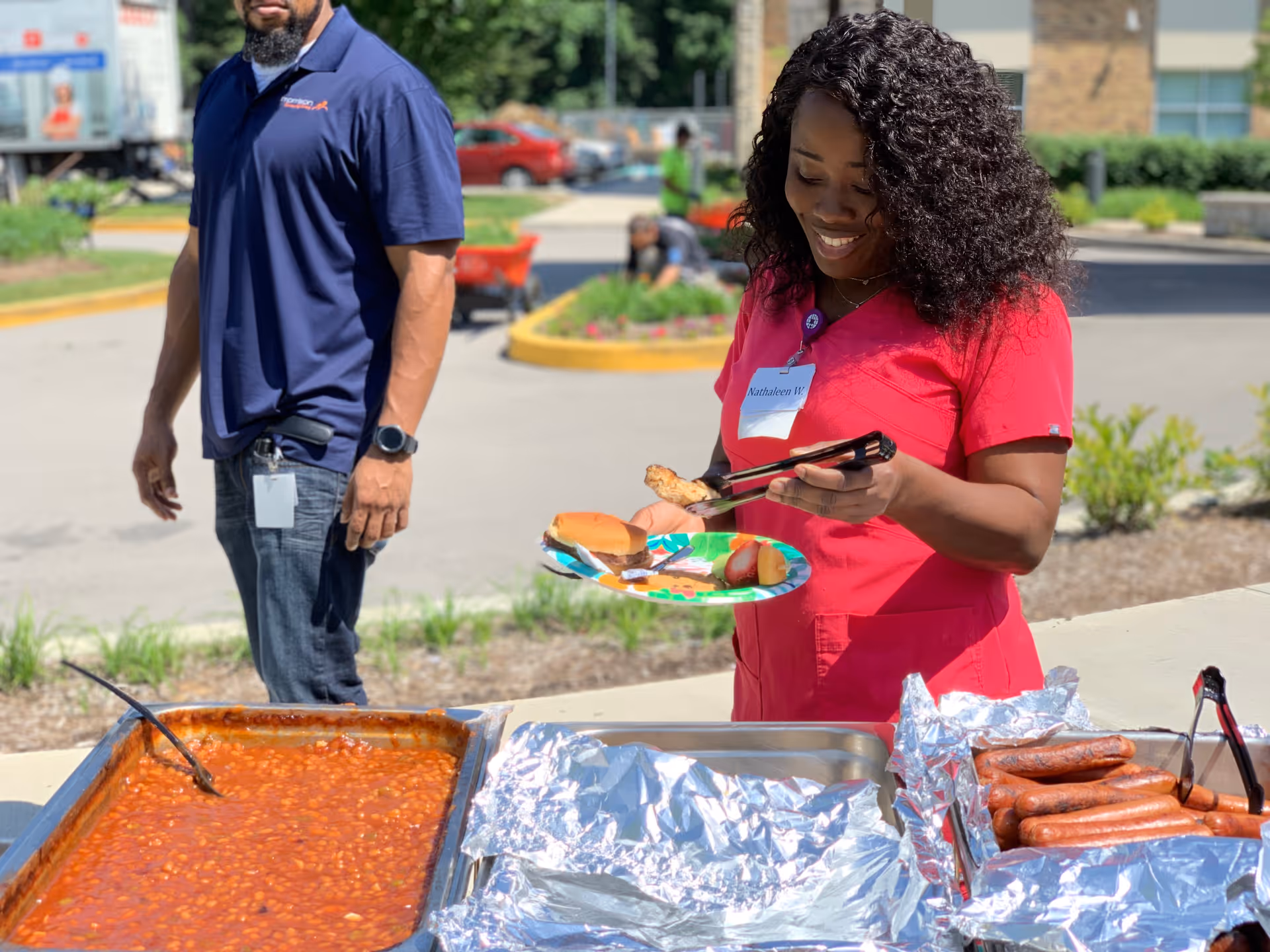 A woman in a red uniform serving food onto a plate outdoors, including a hot dog bun and baked beans, with a man standing nearby in a blue shirt. There are trays of food on a table covered with foil, and a parking lot and greenery in the background.