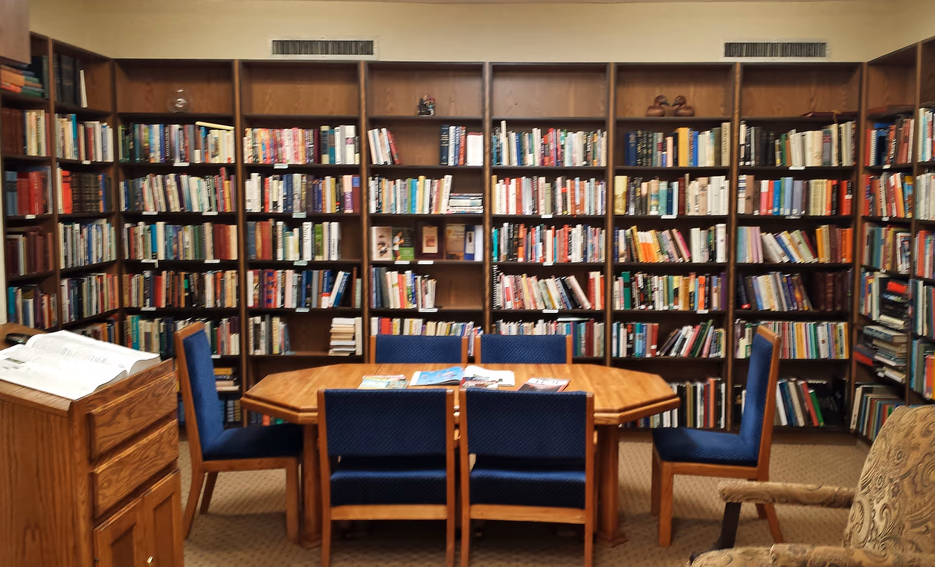 A cozy library room with wooden bookshelves filled with books lining the walls. In the center, there is a wooden table surrounded by six blue upholstered chairs. A wooden lectern with an open book is positioned to the left, and a patterned armchair is partially visible on the right.