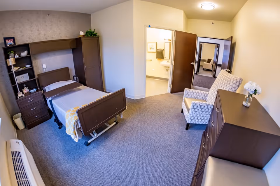 A senior living facility bedroom with a single hospital-style bed, a wooden headboard and footboard, a tall cabinet and shelving unit with decorative items, a patterned armchair, a dresser with a vase of white flowers, and an open door leading to a bathroom and another room with a similar armchair.