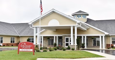 Front exterior view of Orchard Pointe Health Campus building with a covered entrance supported by white columns, an American flag on a flagpole, and a red sign in front that reads 'Orchard Pointe The Residence'. The building has beige siding, a gray roof, and landscaped greenery around the entrance.