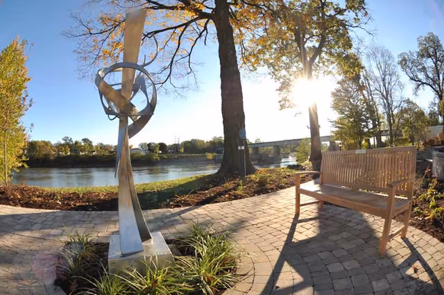 Outdoor paved patio area with a modern metal sculpture on a pedestal surrounded by plants, a wooden bench, large trees, and a river in the background with sunlight shining through the trees.