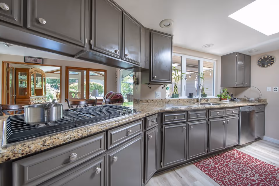 Modern kitchen with gray cabinets, granite countertops, a gas stove with a pot on it, a stainless steel dishwasher, a red patterned rug on the floor, and large windows letting in natural light. There are plants on the countertop and a clock on the wall.