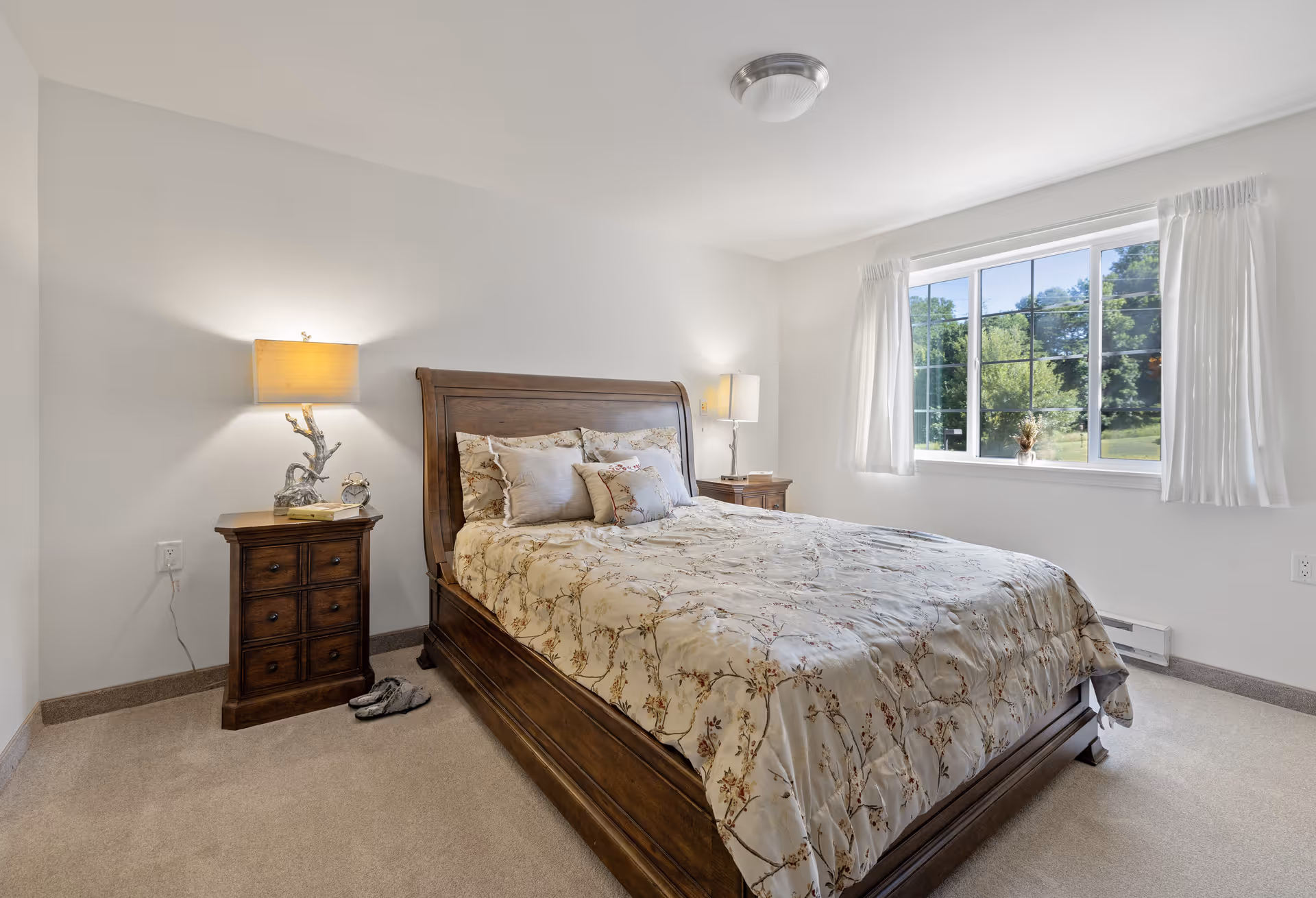 A bright bedroom with a large wooden bed featuring floral bedding and multiple pillows. There are two wooden nightstands on either side of the bed, each with a lamp. A window with white curtains lets in natural light, showing greenery outside. The room has light-colored walls and carpeted flooring.