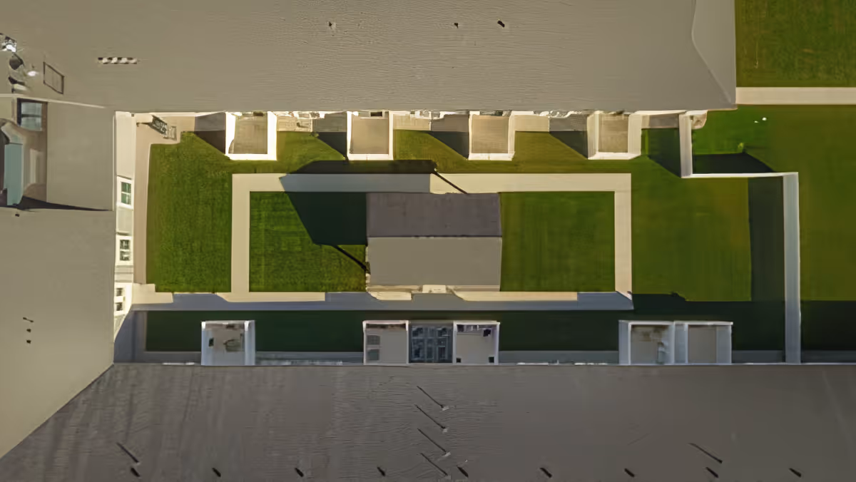Aerial view of a building complex with a central courtyard featuring green grass and paved walkways. The building has a grey roof and white walls surrounding the courtyard area.