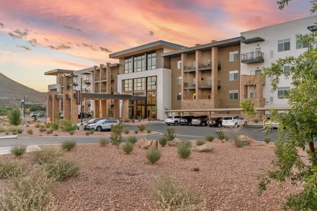 Exterior view of a modern multi-story senior living facility named Legacy Village of St George at sunset, with a landscaped foreground featuring rocks and shrubs, a parking area with several cars, and a mountainous background.