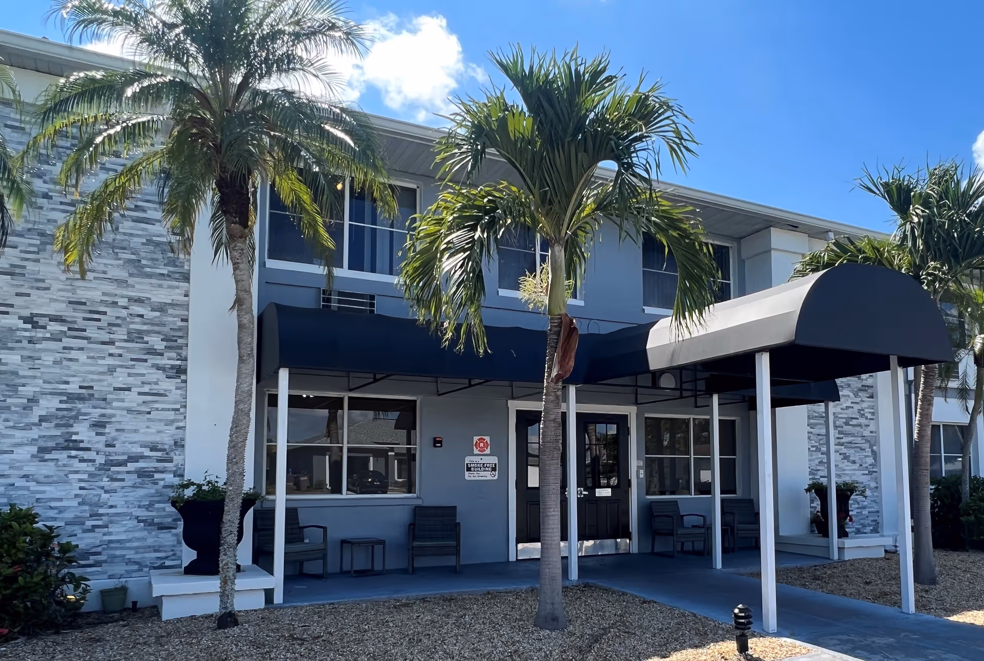 Exterior view of a two-story building with a covered entrance supported by white pillars. The building has a gray and white stone facade with several windows. There are palm trees and some landscaping with gravel in front of the entrance under a clear blue sky.