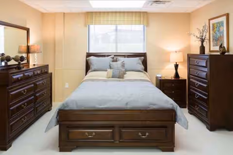 A furnished senior living bedroom with dark wood dressers and a bed by a window.
