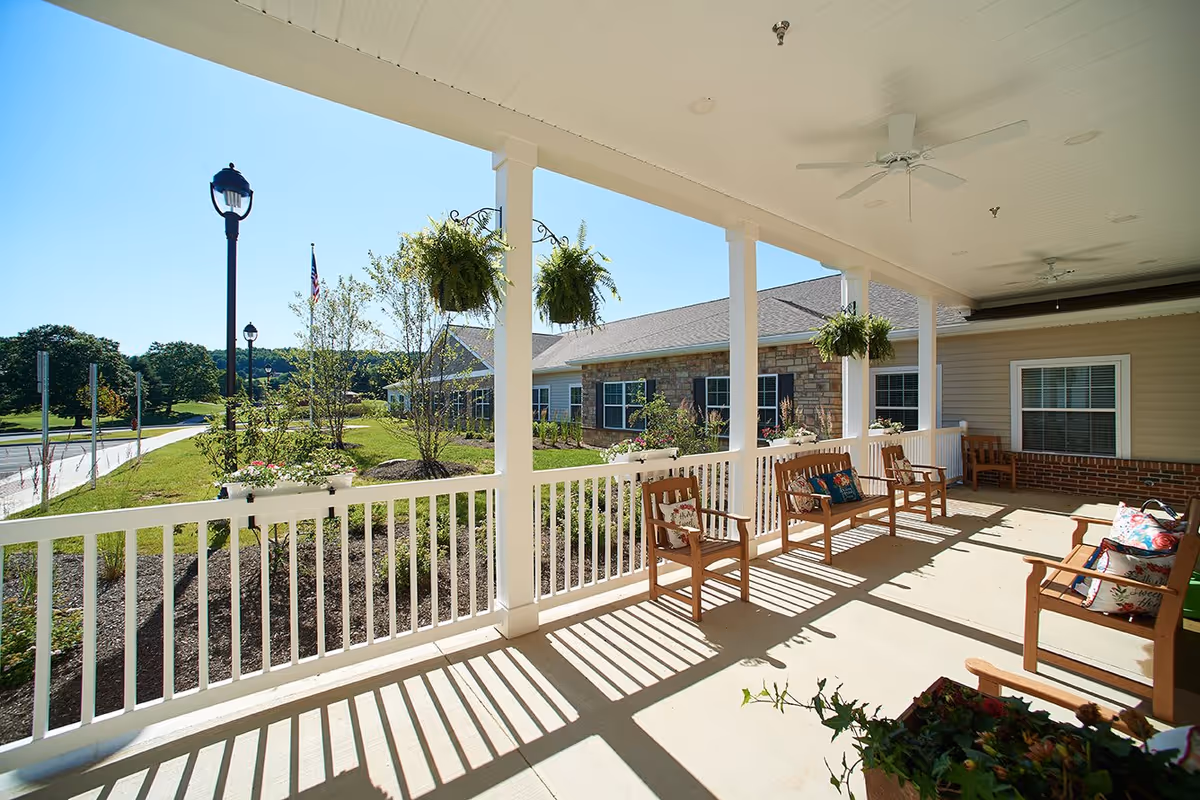 A covered outdoor porch area at Danbury Millersburg with wooden chairs and benches adorned with colorful cushions. White railings and columns support the roof, with hanging green plants adding decoration. The porch overlooks a landscaped garden with trees, flowers, and a clear blue sky.