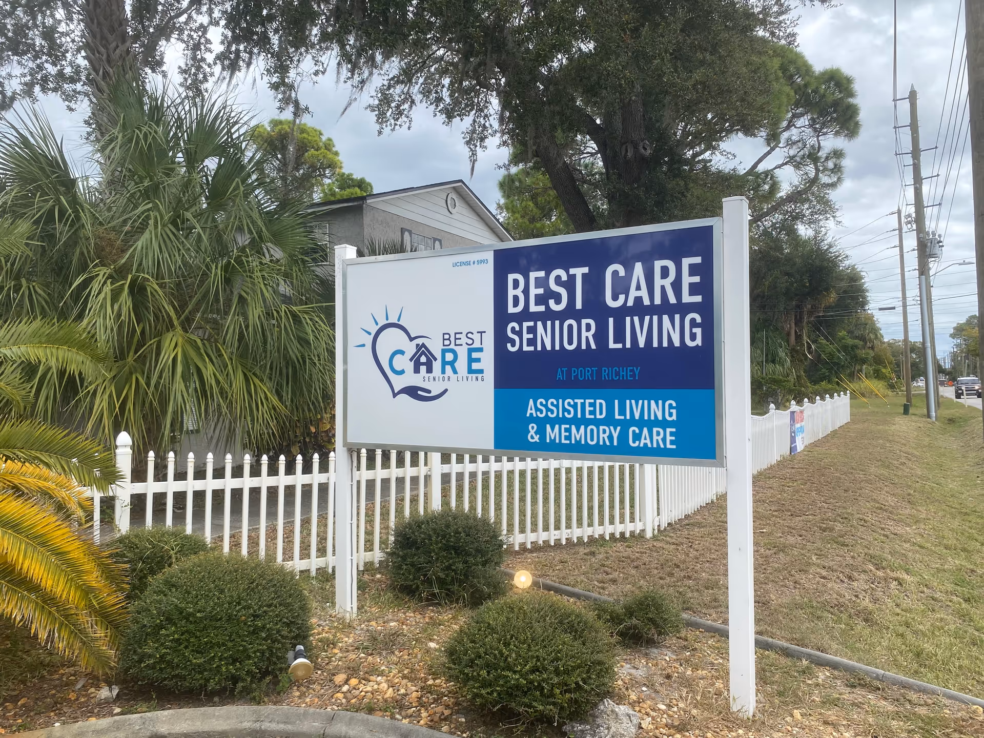 Outdoor sign reading "Best Care Senior Living" in front of a white picket fence with landscaping and trees.