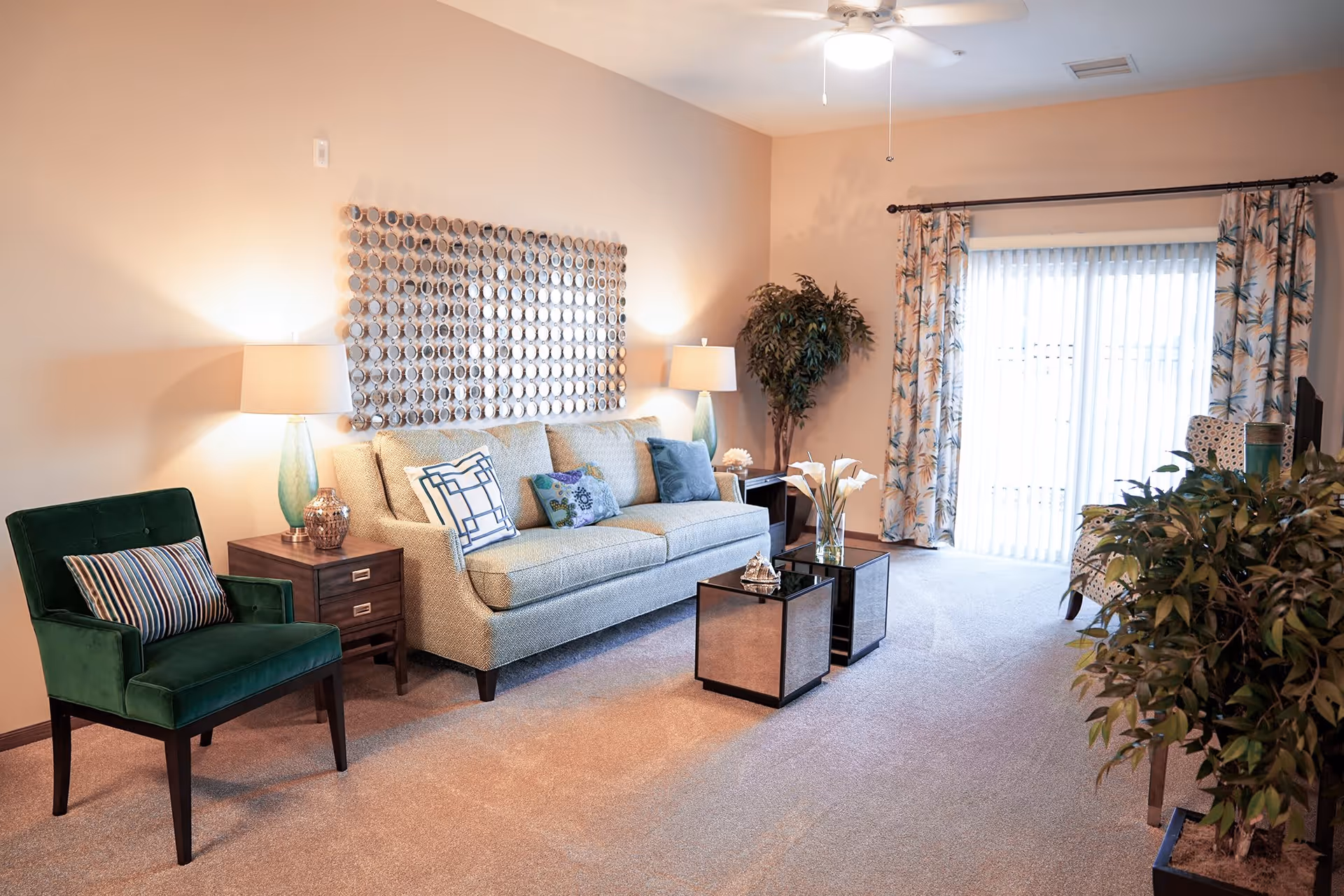 A cozy living room with a beige sofa adorned with decorative pillows, a green upholstered chair with a striped pillow, two side tables with lamps, a decorative wall piece above the sofa, two mirrored cube coffee tables with a vase of white flowers, a large potted plant in the corner, floral curtains covering a sliding glass door, and a ceiling fan with a light.