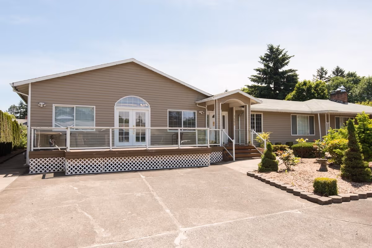 Exterior view of a single-story residential building with beige siding, a small porch with steps, glass railing, and landscaped garden area with shrubs and small trees under a clear sky.