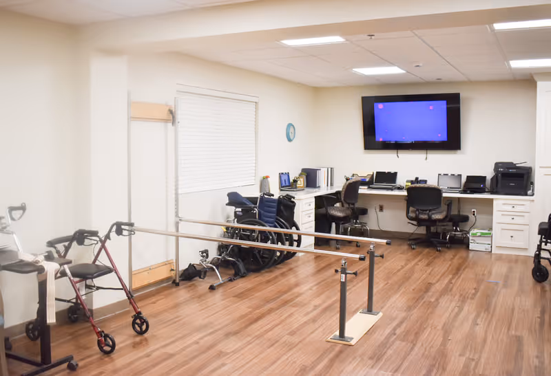 A rehabilitation or therapy room with wooden flooring, parallel bars for walking exercises, several wheelchairs, a walker, and a desk area with office chairs, computers, and a wall-mounted TV screen.