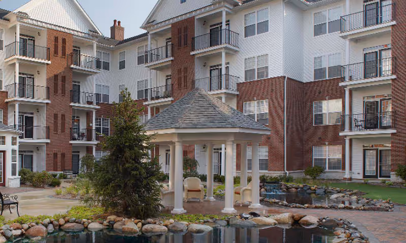 Courtyard of a multi-story brick-and-white senior living building featuring a gazebo, small pond, and balconies.