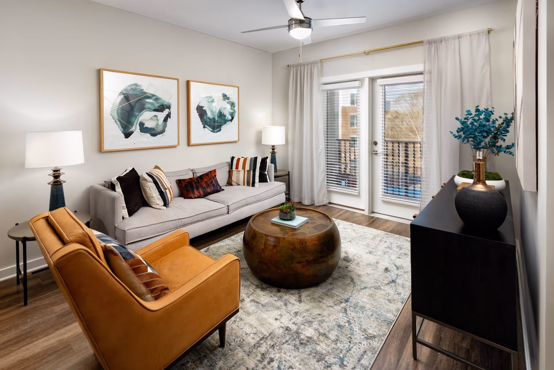 A modern living room with a light gray sofa adorned with multiple colorful pillows, a tan leather armchair, a round wooden coffee table with a small plant on top, two abstract paintings on the wall, two side tables each with a lamp, a black console table with decorative items including a vase with blue-green foliage, and glass doors with blinds leading to a balcony.