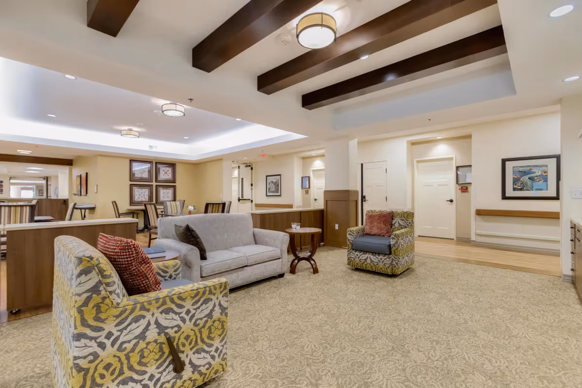 Communal lounge area with a sofa, patterned armchairs, a coffee table, and a dining area under a beamed ceiling.