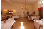 A dining room with several tables covered in white tablecloths, each set with napkins and floral centerpieces. The room features beige walls, a large chandelier hanging from the ceiling, wall sconces with lights, framed artwork, and a mirror. There are upholstered chairs around the tables and a window with a red valance.