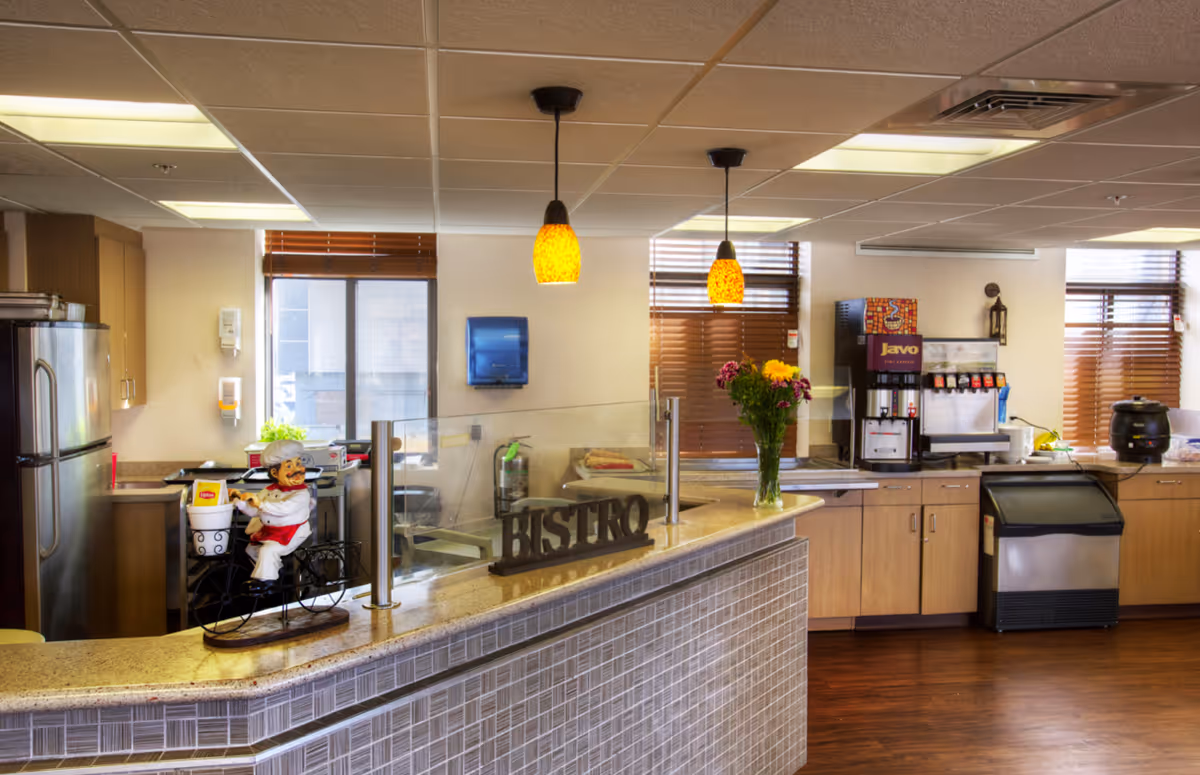 Interior view of a bistro-style kitchen area with a counter displaying a decorative chef figurine on a bicycle, a sign that reads 'BISTRO', and a vase of flowers. The kitchen area includes wooden cabinets, a refrigerator, a coffee machine, and an ice maker. Two pendant lights hang from the ceiling, and windows with wooden blinds allow natural light into the room.