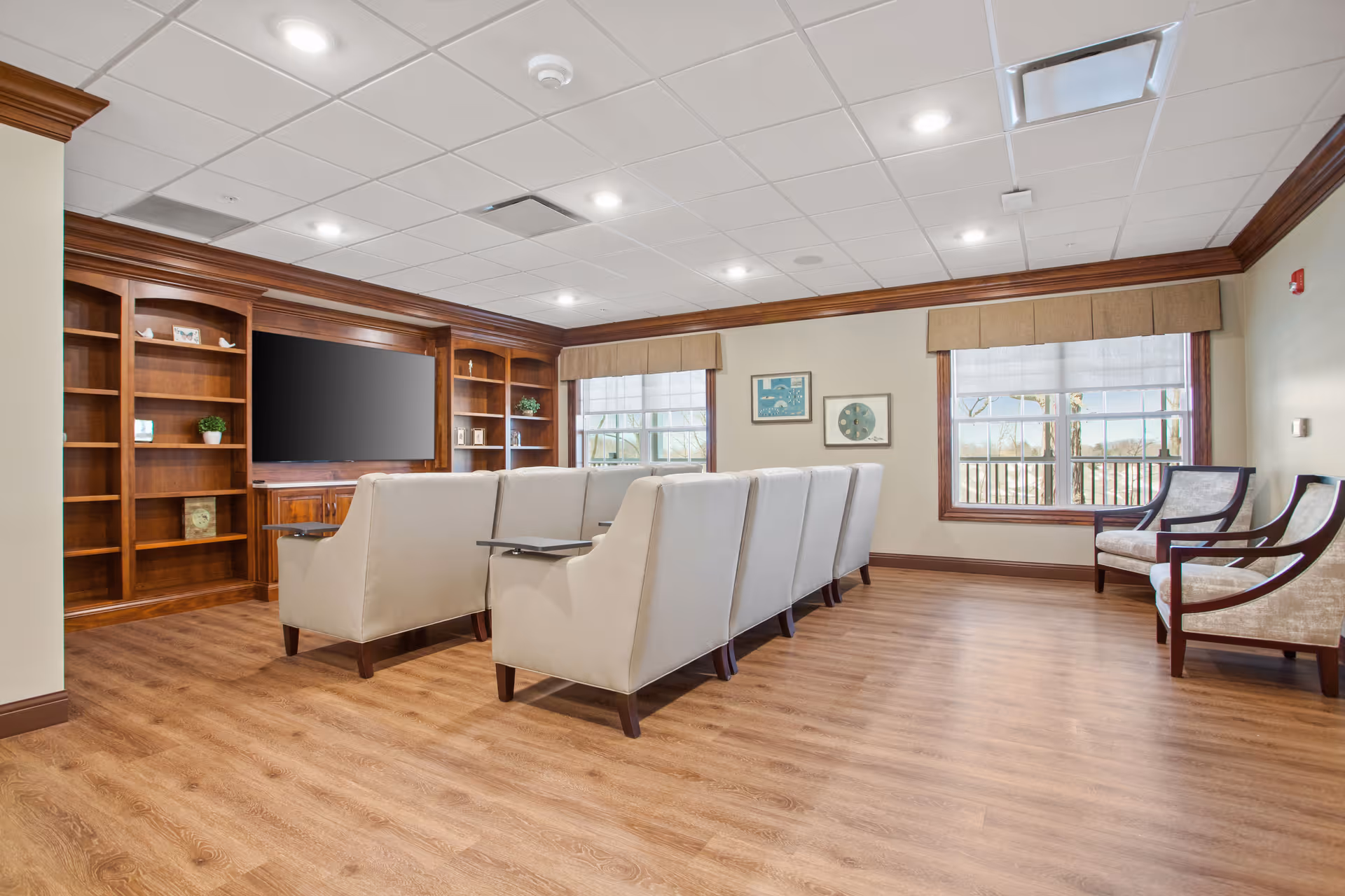 A cozy senior living common area with a row of beige armchairs facing a large flat-screen TV mounted on a wooden built-in shelving unit. The room has wood flooring, two large windows with beige valances, and two additional upholstered chairs near the windows. The walls are decorated with framed artwork.