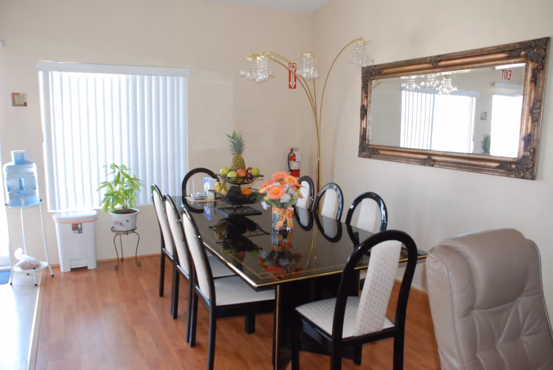 Bright dining room with a glossy black table surrounded by chairs, a large ornate wall mirror, a fruit centerpiece, and a water cooler by a window with vertical blinds.