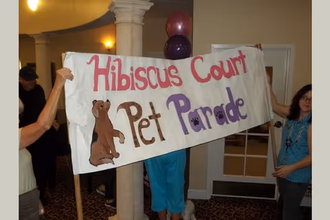 Three people holding a large handmade banner that reads 'Hibiscus Court Pet Parade' with a drawing of a brown dog and purple paw prints. The setting appears to be an indoor common area with carpeted floors, columns, and a door in the background. There are purple and maroon balloons attached to the banner.
